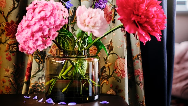 Fresh pink and purple flowers in a green-brown KONSTFULL vase in front of a curtain of NÄSSELKLOCKA textile.