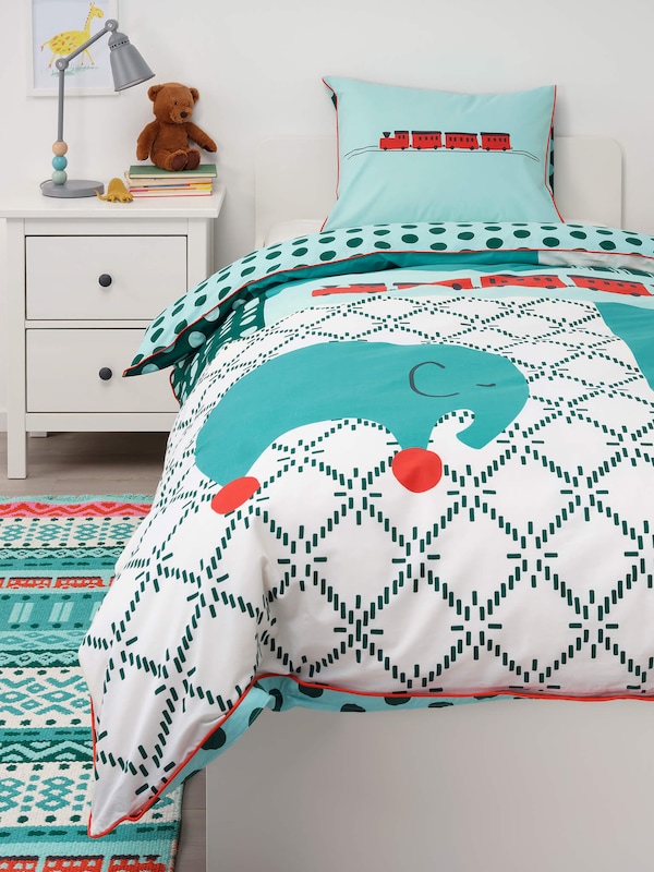 Kids bedding with a colorful, fun design next to a decorative rug & a nightstand with a teddy bear. 