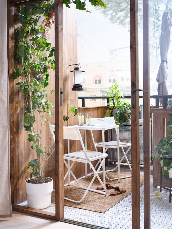 Open doors leading to a leafy balcony which has a white table and two chairs on it, with a lantern on the wall above.