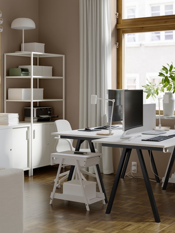 A living room with an office chair, lamps and two TROTTEN desks placed against each other with a noticeboard in the middle.