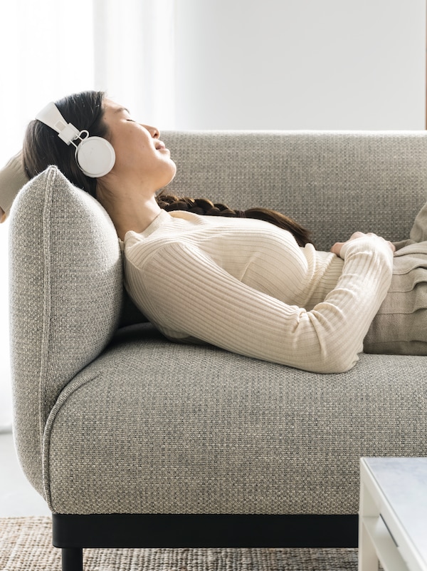 A woman with white headphones is reclining on a light grey ÄPPLARYD sofa with her head and an arm up on the armrest.