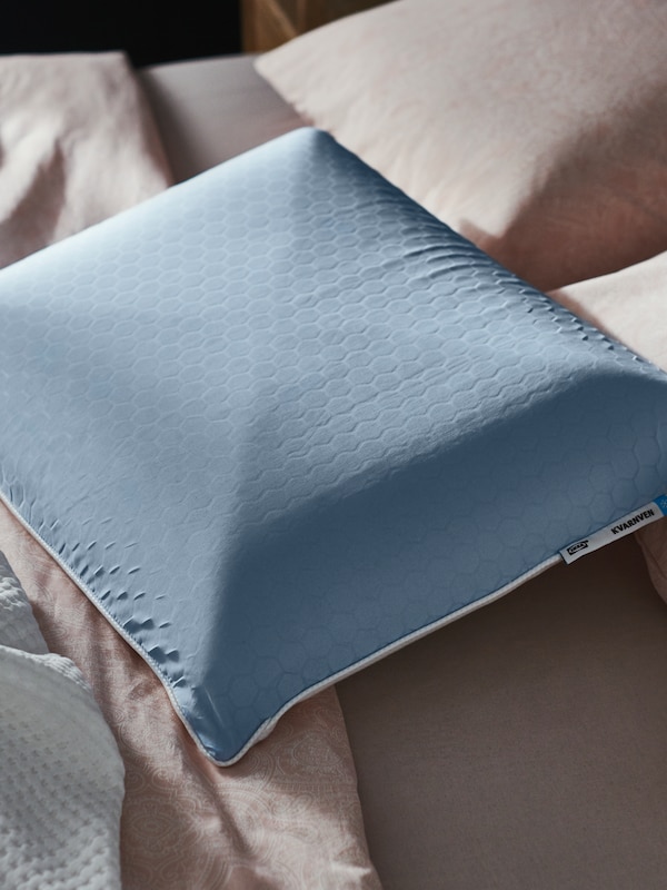 A KVARNVEN ergonomic pillow, which has a cooling blue side with a honeycomb pattern, lies on top of a bed.