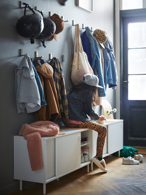 A hallway with a child sitting on a white MACKAPÄR bench with storage, with clothes and helmets on hooks on the wall.