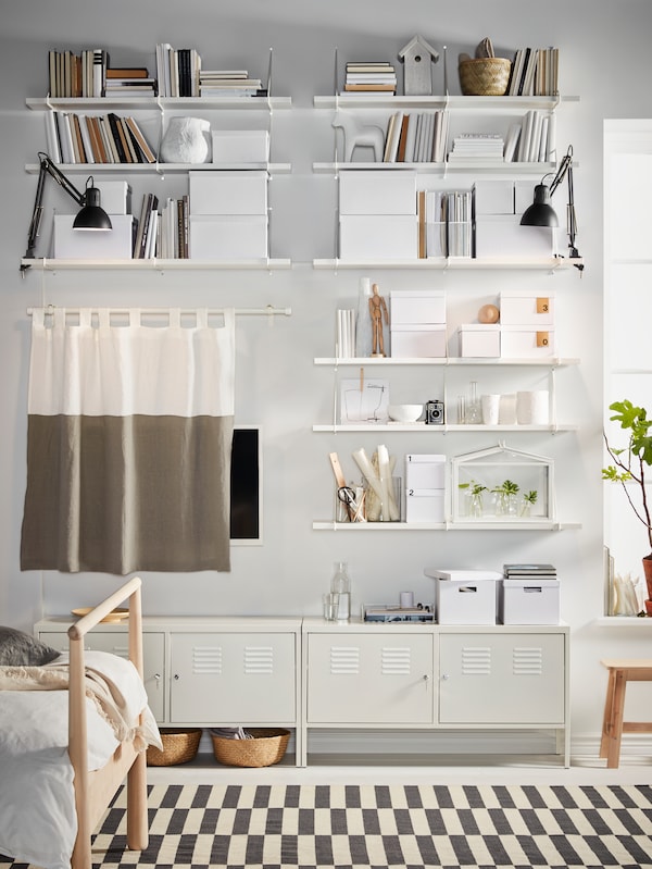 A wall with BERGSHULT/PERSHULT wall shelf combinations full of books and boxes, a curtain hiding a TV and two cabinets below.