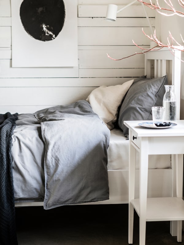 A white stain HEMNES bedside table stands beside a white stain HEMNES bed with grey ÄNGSLILJA bed linen.