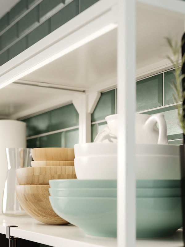 A white shelving unit holding bowls and dishes in wood and green and white porcelain, plus integrated lighting.