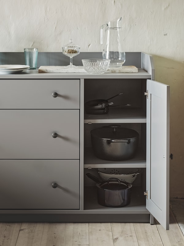 A grey HAUGA sideboard with an open door showing an OUMBÄRLIG saucepan and a VARDAGEN pot with lid. A glass jug is on top.