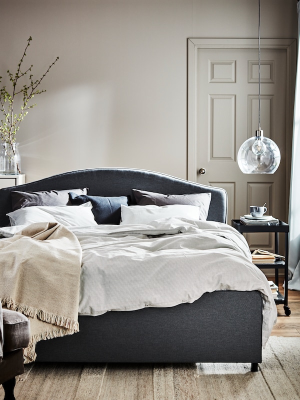 A JAKOBSBYN pendant lamp hangs in a bedroom near a grey HAUGA upholstered bed with BERGPALM bed linen.