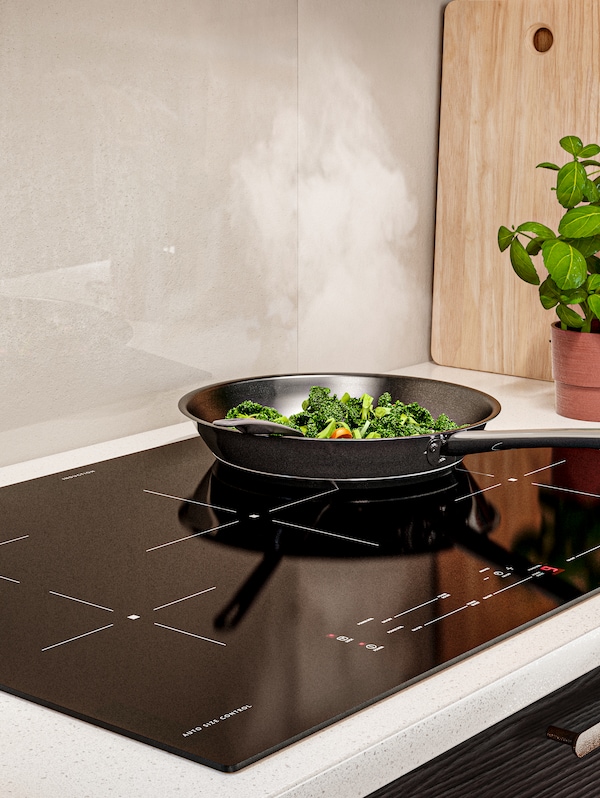A frying pan is stir-frying vegetables on a SÄRKLASSIG induction cooktops. A pot of herbs is beside it on a white worktop.