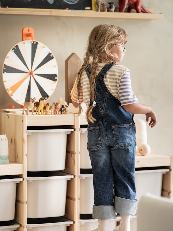 A young child stands beside a TROFAST storage combination which has a LUSTIGT wheel of fortune and toy animals on top.