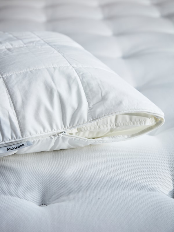 A pillow which is inside a partly zipped up ÄNGSKORN pillow protector lies on top of a mattress.