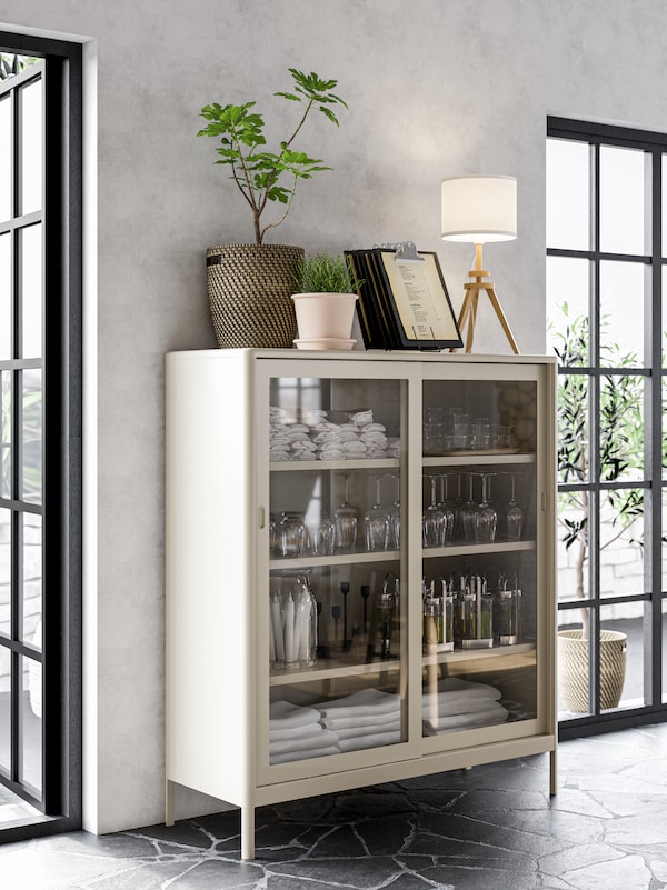 A beige cabinet with sliding glass doors with napkins, glasses and candles stored neatly in it.