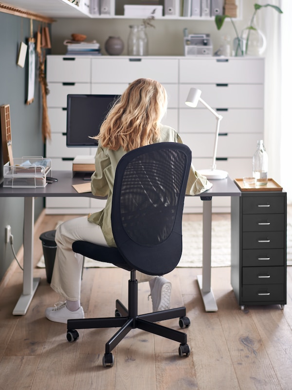 A woman sitting in a black FLINTAN office chair by a grey sit/stand RODULF desk with a computer, work lamp and drawer unit.