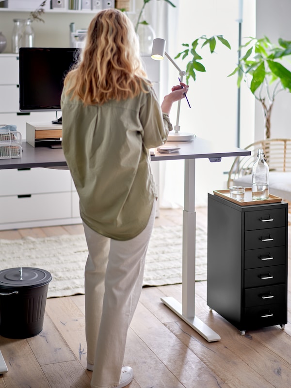 A home office with a woman standing at a grey sit/stand RODULF desk with a computer, a work lamp, and a drawer unit.