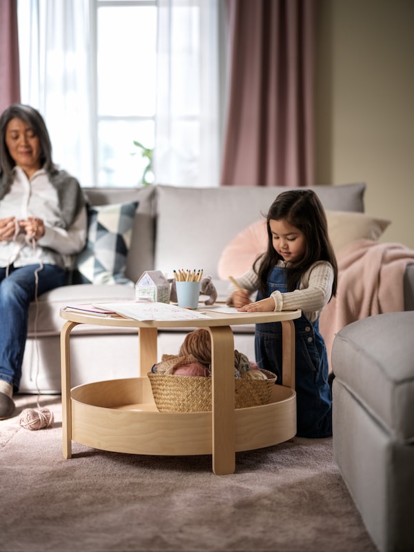 A living room where a girl draws at a BORGEBY birch-veneer coffee table, and a woman crochets in a light-grey sofa.