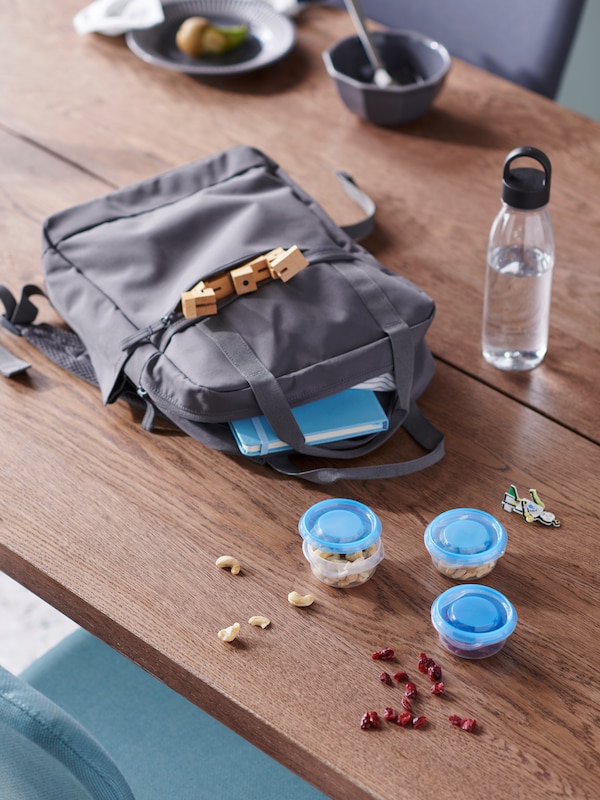 A grey STARTTID backpack, a filled IKEA 365+ water bottle and snack-filled PRUTA containers lying on a wooden tabletop.