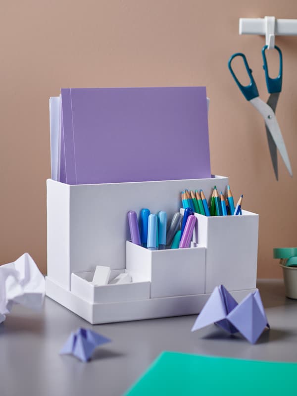 A desktop with colorful folded paper, scissors hanging from a SUNNERSTA rail, and pens and paper in a TJENA organizer.