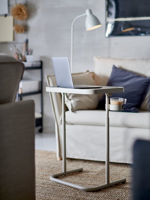 A laptop on a beige BJÖRKÅSEN laptop stand, a jute rug, an armchair with cushions and a white floor/reading lamp.