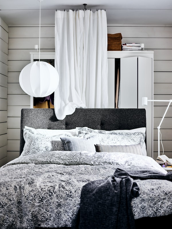 A dark grey IDANÄS upholstered bed with white/grey ÄNGSKLOCKA bed linen stands in front of a white IDANÄS wardrobe.