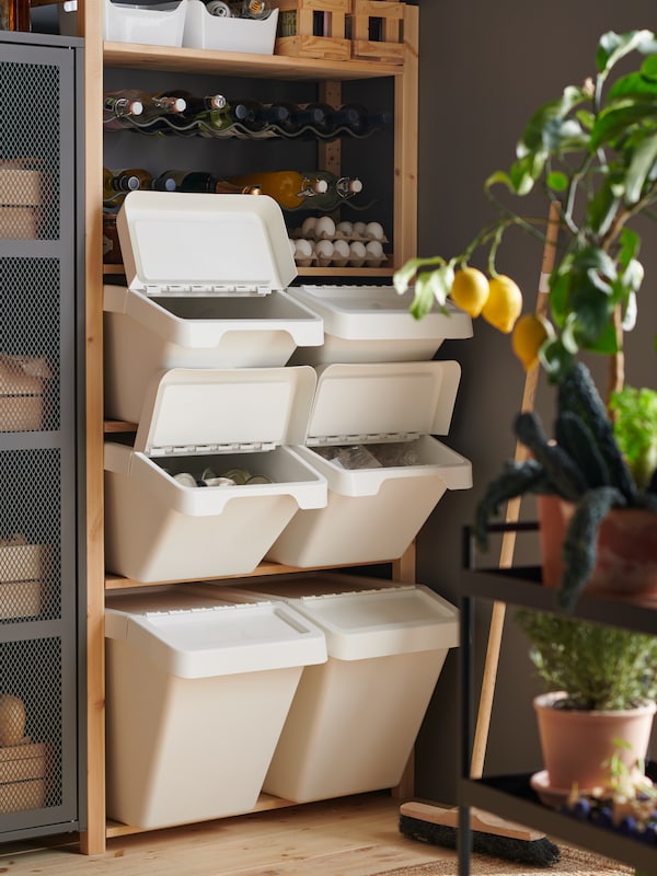 A corner of a room with an IVAR shelving unit filled with different-size white SORTERA waste bins beneath a bottle rack.