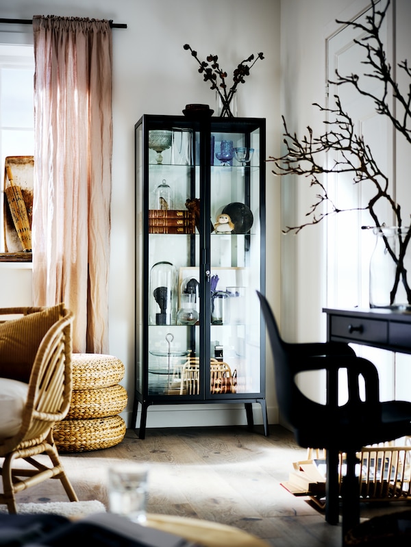 A black MILSBO cabinet with glass doors filled with an assortment of objects, in the corner of a bright living room.