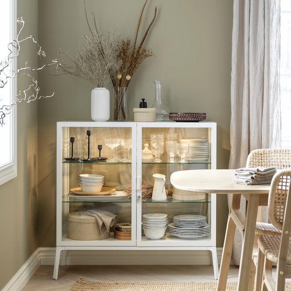 A white glass-door cabinet with integrated lighting stands in the corner of a dining room and is filled with tableware.