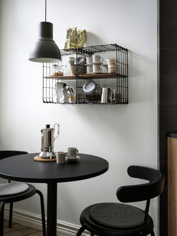 Two GULLHULT wall shelves holding coffee and tea accessories hang above a small, round table and two chairs.