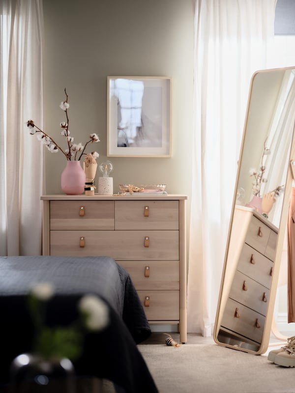 A bedroom where a BJÖRKSNÄS birch chest of drawers with decorations on top stands next to an IKORNNES ash standing mirror.