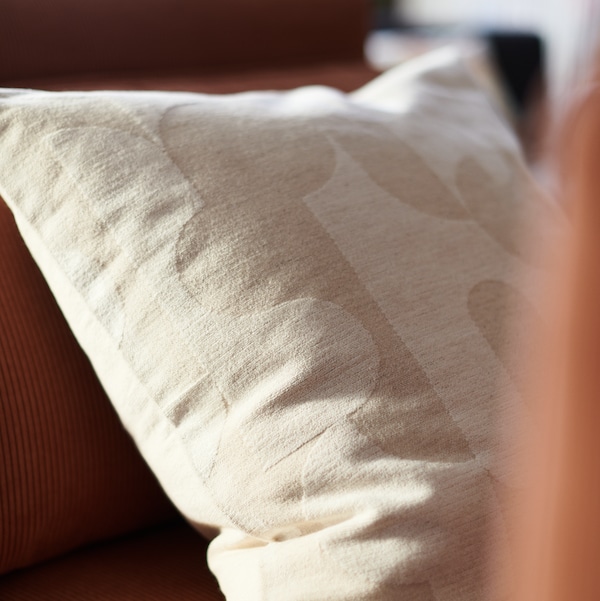 A cushion which has a beige RÖDASK cushion cover lies at an angle on a sofa.