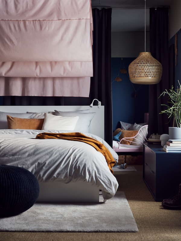 A bamboo MISTERHULT pendant lamp hangs near a white MALM bed which stands in front of a room divider made of curtains.