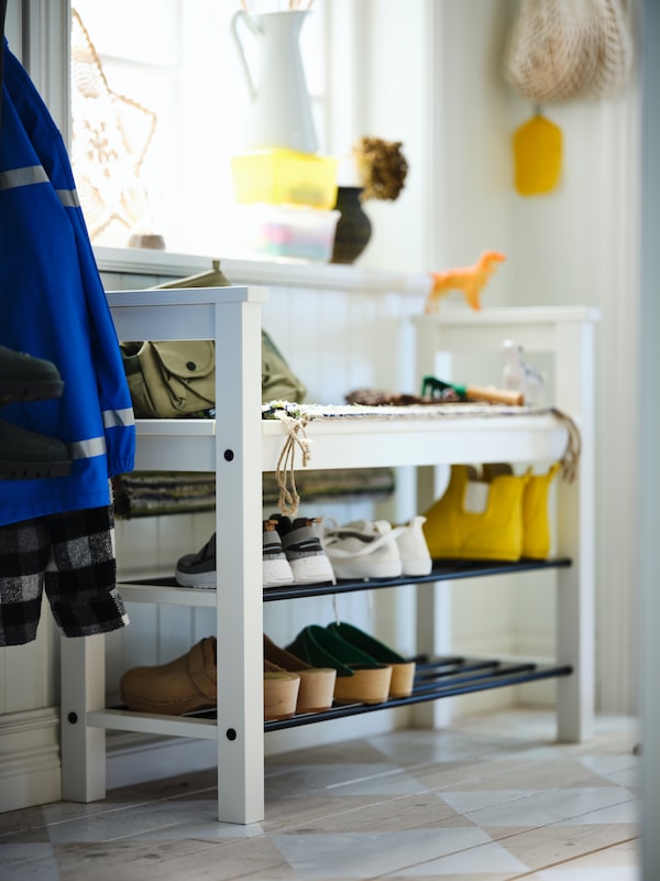 A HEMNES bench with shoe storage holding shoes and a bag stands in a hallway with children’s clothing hanging beside it.