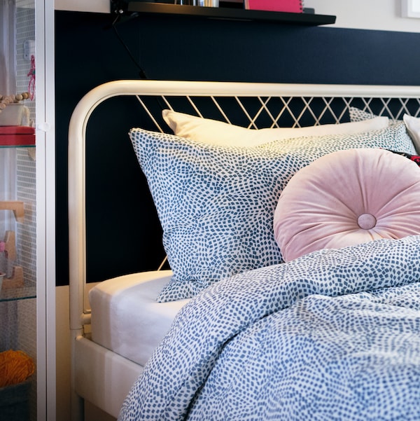 The head of a white NESTTUN bed with white/blue TRÄDKRASSULA bed linen and a light pink KRANSBORRE cushion.