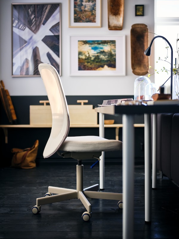 A beige FLINTAN office chair standing by a desk with a dark-grey LAGKAPTEN tabletop in a living room with dark wooden floor.