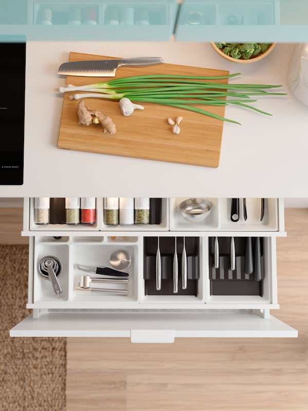 A white countertop with a chopping board on it with a pulled-out drawer below, showing neatly organised tools and utensils.