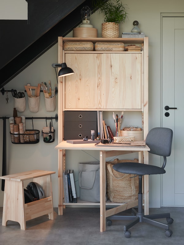 A pine storage unit with a desk under a flight of stairs. Containers on a wall hold tools. A clamp spotlight is on the desk.