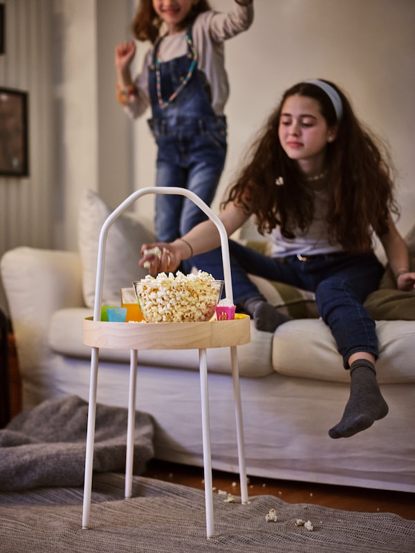 One girl standing on a sofa, while another sits and grabs popcorn from a bowl on a white BURVIK side table.
