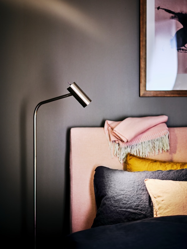 A VIRRMO floor/reading lamp stands beside a pale pink IDANÄS upholstered bed with DYTÅG bed linen and extra cushions.