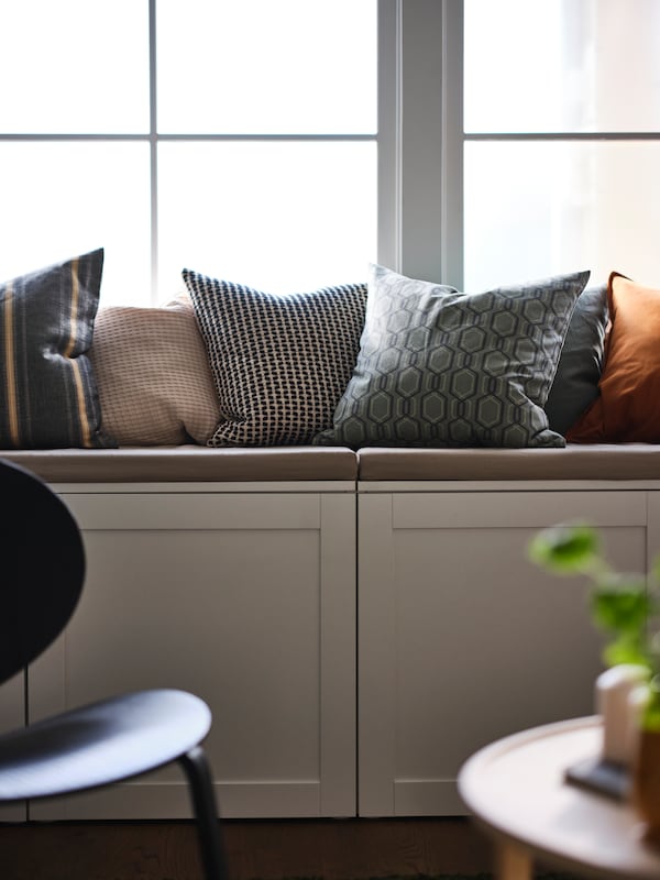 A storage bench with a lot of cushions on top, including one in a JÄTTEPOPPEL cushion cover, stands under a window.