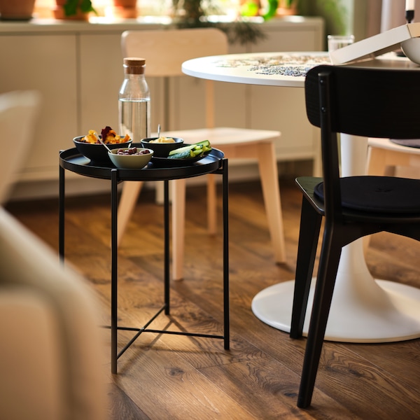 A black GLADOM tray table covered in snacks next to a white DOCKSTA table with two LISABO chairs in black and ash.