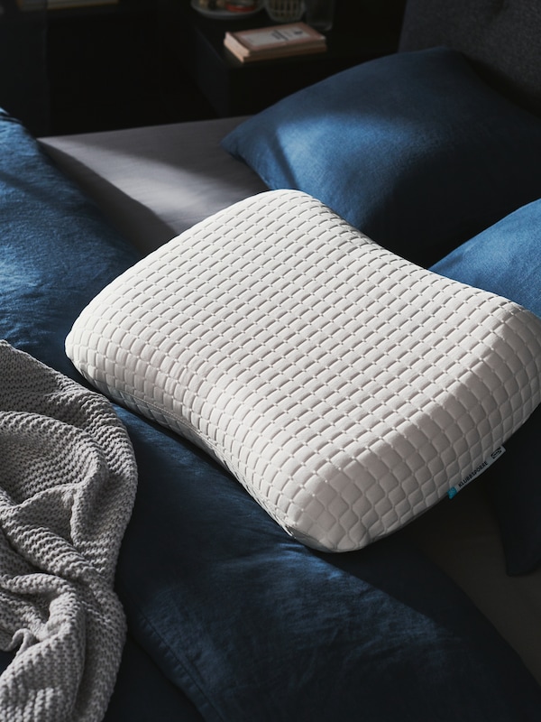A KLUBBSPORRE ergonomic pillow lies next to a grey throw on top of a bed with dark blue bed linen.