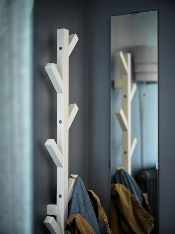 A coat hangs on a branch-shaped, wall-mounted, white TJUSIG hanger next to a mirror in a corner of a room with grey walls.