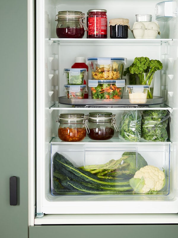 An open fridge filled with vegetables, jars and food containers, some of which are placed on a SNURRAD storage turntable.
