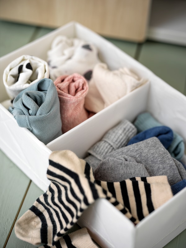 A white STUK box with compartments containing a mix of clothes sits on a floor. A striped sock lies on one edge of the box.