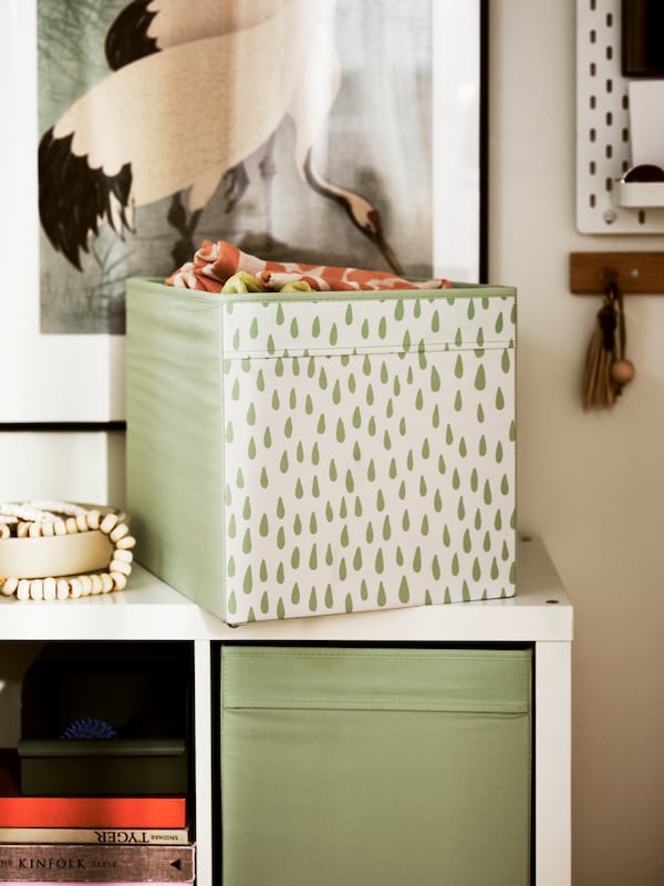 A DRÖNA box stands on top of a KALLAX shelving unit in a hallway. Another DRÖNA box sits on a shelf in the shelving unit.