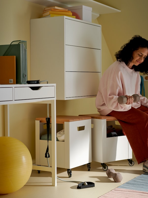 A woman holding weights sits on a TROTTEN storage unit on castors in a workspace at home.