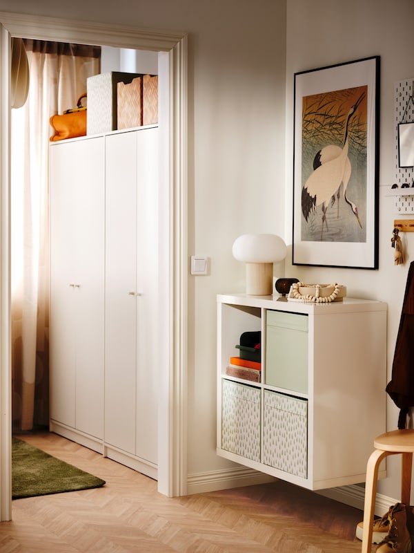 A KALLAX shelving unit with DRÖNA boxes is mounted to the wall in a hallway near a room with two white wardrobes.