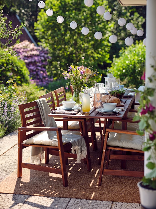 ÄPPLARÖ wooden chairs and a table laid for a meal on top of a rug on a leafy terrace with an LED lighting chain above.