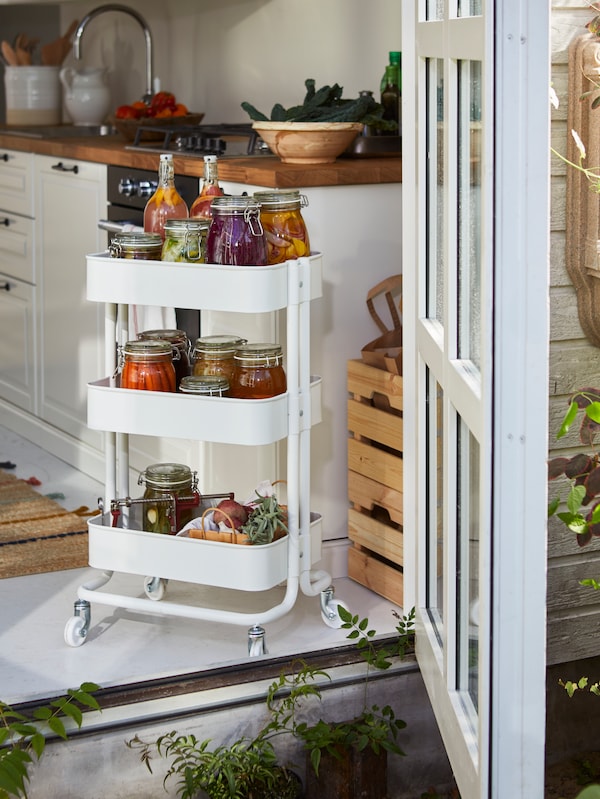 A selection of glass jars contain pickled vegetables and other colorful foods, all filling a RÅSKOG cart on wheels.