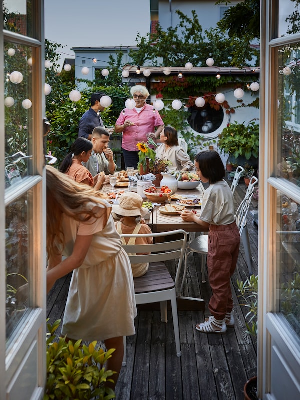 A group of adults and children, friends and family gather around a NORRMANSÖ outdoor table and chairs.