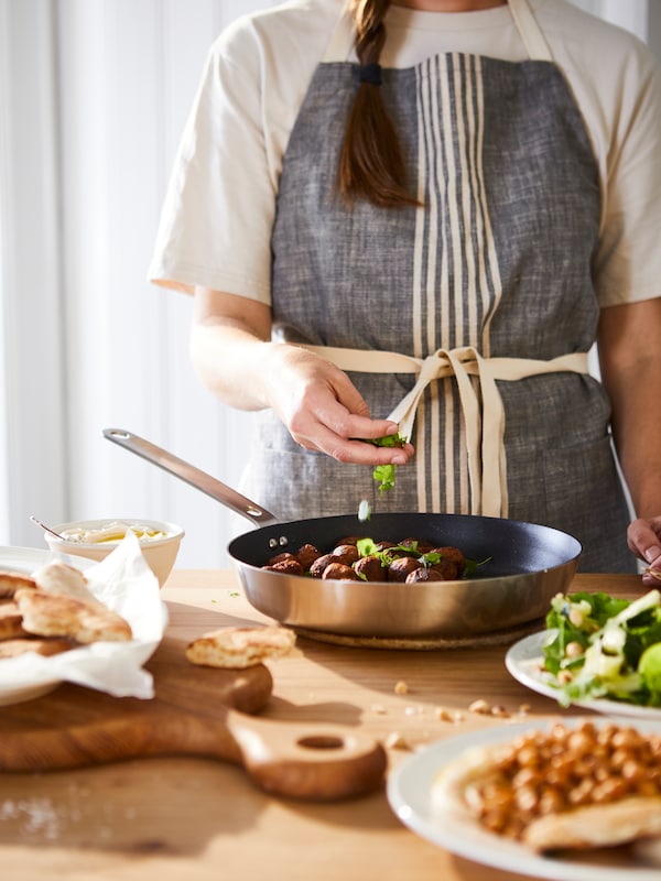 A person stands in a kitchen, cooking with a HEMKOMST frying pan and tossing ingredients in.